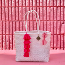 Load image into Gallery viewer, There is a white handmade from recycled plastic large tote sitting on a pink floor. On the large tote hang one pair of pink heart shaped glasses and one large tassel. Behind the tote is a pink brick wall.