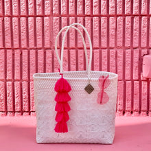 Load image into Gallery viewer, There is a white handmade from recycled plastic large tote sitting on a pink floor. On the large tote hang one pair of pink heart shaped glasses and one large tassel. Behind the tote is a pink brick wall. 
